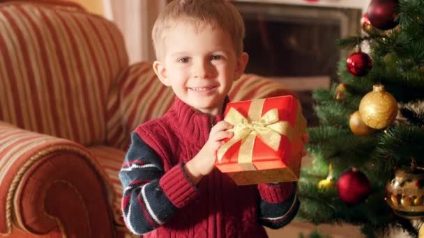 4k video de niño riendo feliz con regalo de Navidad en una hermosa caja de pie en el árbol de Navidad y mirando en la cámara. Niño recibiendo regalos y regalos de Santa Claus en las vacaciones de invierno y — Vídeo de stock