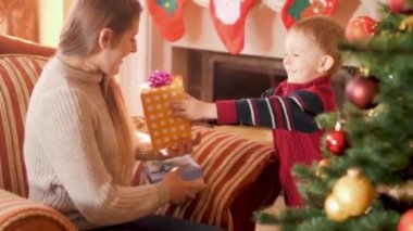 Mutlu gülümseyen küçük çocuk Noel ağacının yanında annesine kurdele ile Noel hediye kutusu veren 4k video. Kış tatillerinde Noel Baba'dan hediye ve hediye alan çocuk ve