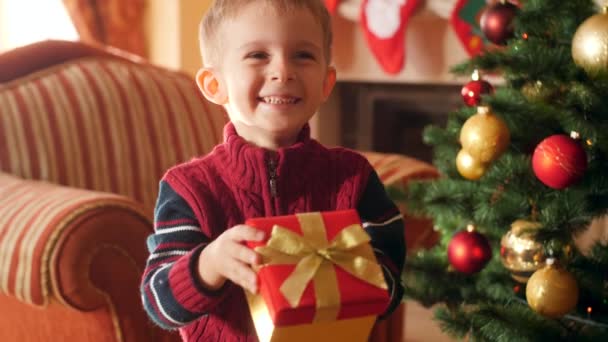 4k vídeo de feliz sorrindo menino segurando presente de Natal na caixa e mostrando seu presente na câmera. Criança recebendo presentes e presentes do Papai Noel em férias de inverno e celebrações — Vídeo de Stock