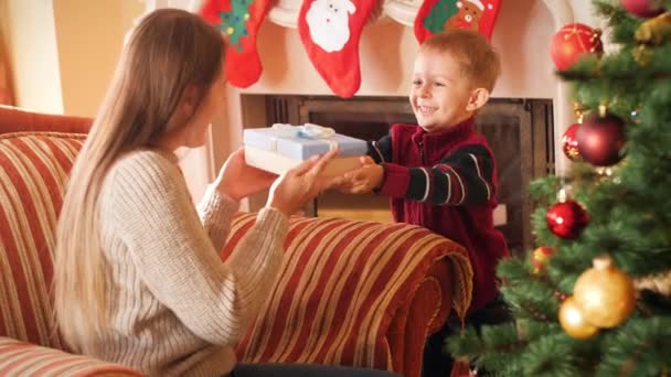 Wideo 4K uśmiechniętego małego chłopca dając Boże Narodzenie prezent dla matki. Rodzina dając i otrzymując nowy rok prezentuje w salonie obok choinki. — Wideo stockowe