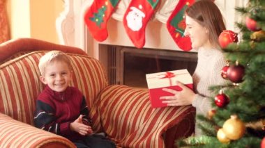 Genç anne güzel Noel ağacı yanında koltukta oturan küçük oğluna Noel hediyesi veren 4k video. Noel ağacının yanındaki oturma odasında aile hediye ve yeni yıl hediyesi alma.