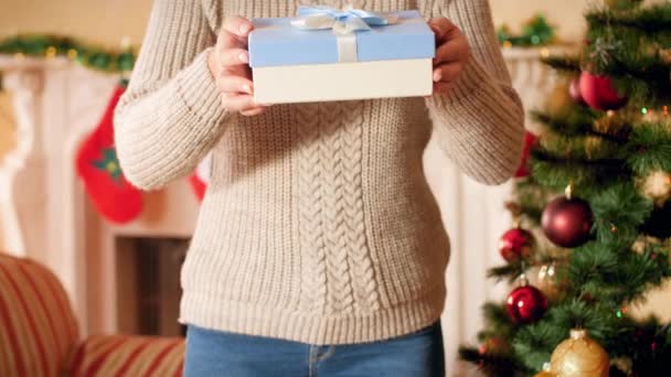 4k footage of young woman in wool sweater standing next to Christmas tree and fireplace and showing Christmas present box with ribbon and bow. — Stock Video