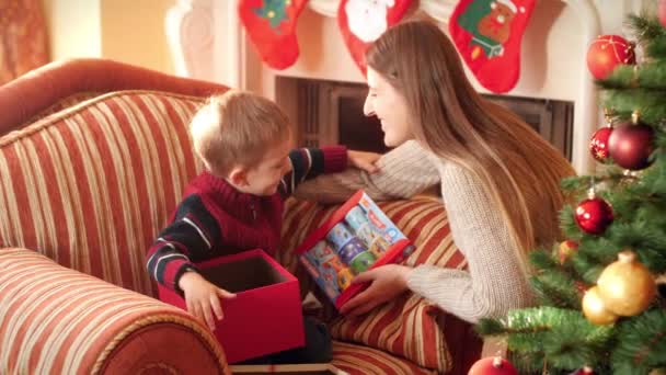 Filmagem 4k da jovem mãe beijando seu filhinho depois de dar-lhe presente de Natal. Família dando e recebendo presentes de Ano Novo na sala de estar ao lado da árvore de Natal . — Vídeo de Stock