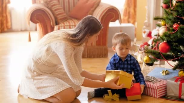 크리스마스 트리 아래 그녀의 작은 아들과 함께 앉아 크리스마스 선물과 선물을 여는 행복한 젊은 어머니의 4K 비디오. 겨울 방학 및 축하 에 가족 기부 및 수령 선물. — 비디오