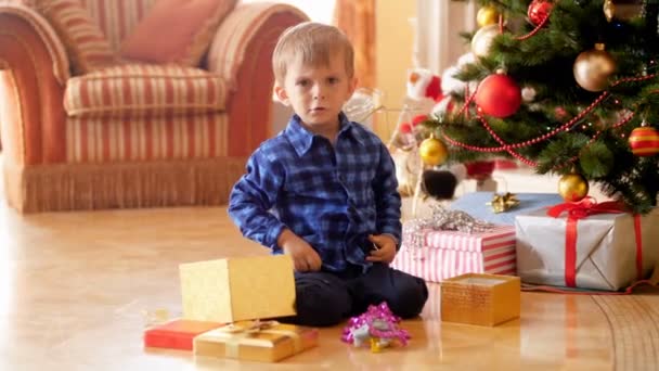 4k videó dühös fiú ül a karácsonyfa alatt, és kiabál, mert a nem kívánt ajándékokat, és jelen ő rendelte a Mikulás. — Stock videók