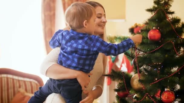 Filmagem 4k de feliz sorridente jovem mãe com o filho pendurado bugigangas e brinquedos na árvore de Natal na sala de estar. Casa de decoração familiar em férias de inverno e celebrações . — Vídeo de Stock