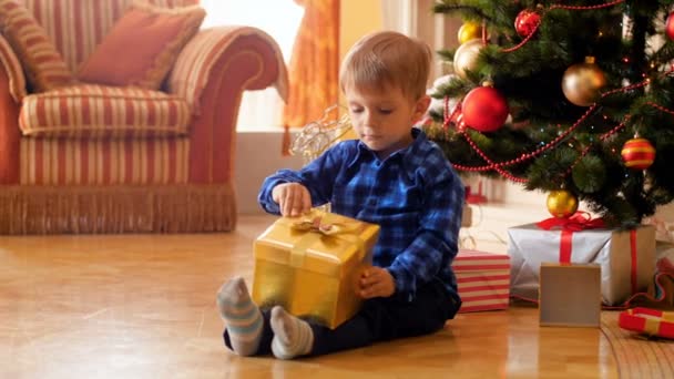 4k video of little cute boy sitting on the floor next to Christmas tree and opening box with present from Santa Claus. Family having good time and fun on winter holidays and celebrations. — 비디오
