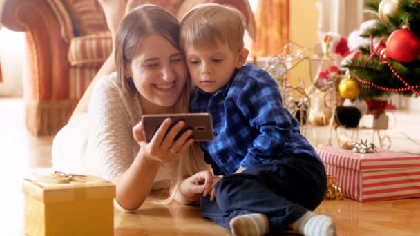 4k video med glada leende fmaily liggande på golvet under julgranen och med hjälp av smartphone. Familjen har roligt på vinterlovet och firandet. — Stockvideo