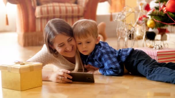 4k-beelden van kleine jongen met jonge moeder liggend op de vloer in de woonkamer en het bekijken van cartoons op de smartphone. Familie met goede tijd en plezier op Winter vakantie en feesten. — Stockvideo