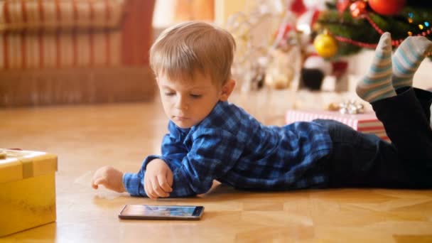 4k video of cute little boy lying on floor next to Christmas tree and watching cartoons on smartphone. Child having good time and fun on winter holidays and celebrations. — Stok video