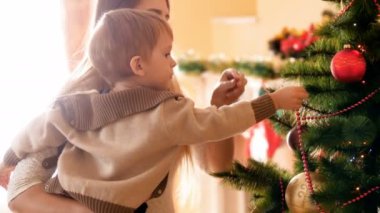 Küçük çocuğun annesine noel ağacını süsleyen süs eşyaları ve oyuncaklarla yardım ederken çekilmiş 4k görüntüleri. Kış tatillerinde ve kutlamalarda aile ev hazırlama ve dekorasyon.