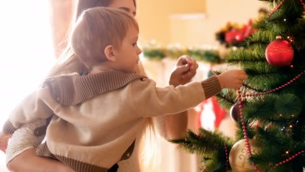 Imágenes de 4k del niño ayudando a su madre a decorar el árbol de Navidad con chucherías y juguetes. Familia preparando y decorando la casa en las vacaciones de invierno y celebraciones . — Vídeos de Stock