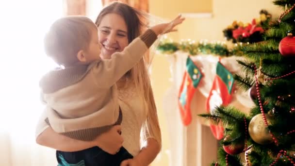 4k video de la feliz madre joven sosteniendo y abrazando a su pequeño hijo junto al árbol de Navidad en la sala de estar. Familia pasar un buen rato y diversión en las vacaciones de invierno y celebraciones . — Vídeos de Stock