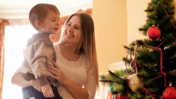 4k metraje de madre feliz msiling con su pequeño hijo de pie y mirando el árbol de Navidad descongelado en casa. Familia pasar un buen rato y diversión en las vacaciones de invierno y celebraciones . — Vídeos de Stock