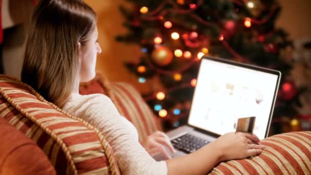 4k视频的年轻女子放松在客厅的大扶手椅在发光的圣诞树旁边，手里拿着信用卡和网上购物。网上购物和电子商务的概念镜头. — 图库视频影像