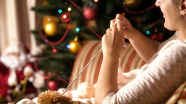 Closeup 4k footage of young woman sitting in armchair next to glowing christmas tree and knitting wool sweater for her children. People relaxing on winter holidays and celebrations at home — Stock Video