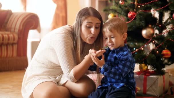 Vídeo em câmera lenta de mãe sorridente feliz com seu filho soprando confete espumante da mão enquanto se senta sob a árvore de Natal na sala de estar. Família se divertindo e se divertindo nas férias de inverno — Vídeo de Stock