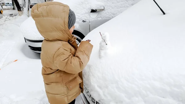 雪が降った後に雪に覆われた車を掃除する少年の画像.寒い冬の朝に — ストック写真