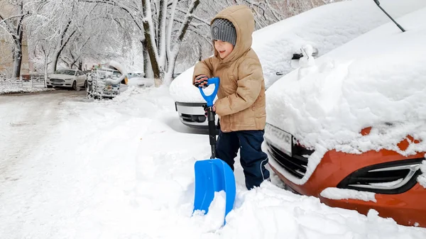 Närbild foto av leende söta småbarn pojke i beige jacka och grå hatt hjälper till att städa upp snötäckta röd bil efter snöstorm med stor blå spade — Stockfoto