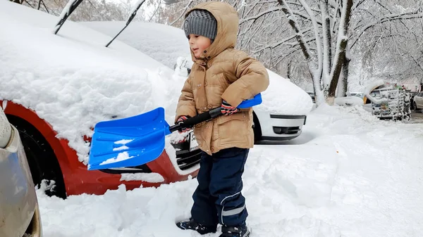 Imagen de niño con chaqueta ayudando a limpiar el coche cubierto de nieve después de ventisca usando una gran pala azul —  Fotos de Stock