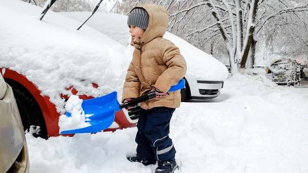 Foto do menino sorridente na jaqueta ajudando a limpar o carro coberto de neve após a nevasca usando uma grande pá azul — Fotografia de Stock