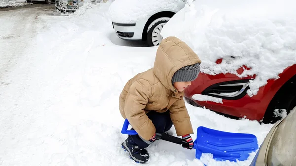 Zbliżenie strzał uśmiechnięty uroczy chłopiec w marynarce i kapeluszu pomaga oczyścić śnieg pokryte samochód po zamieć za pomocą Big Blue łopata — Zdjęcie stockowe