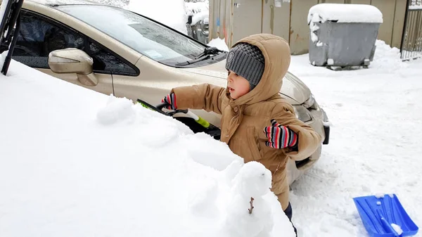 Foto de niño ayudando a limpiar el coche cubierto de nieve después de ventisca usando gran pala azul —  Fotos de Stock