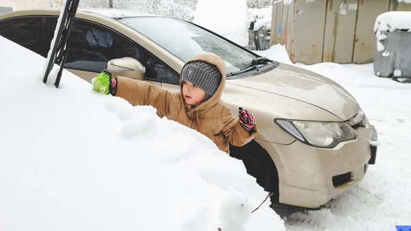 Foto de primer plano del niño feliz ayudando a limpiar y desenterrar el coche cubierto de nieve después de la caída de nieve —  Fotos de Stock