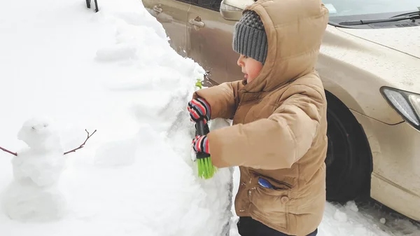Feche a foto do menino na jaqueta limpe o carro coberto de neve após a tempestade de neve usando o pincel grande. Na manhã fria do inverno — Fotografia de Stock