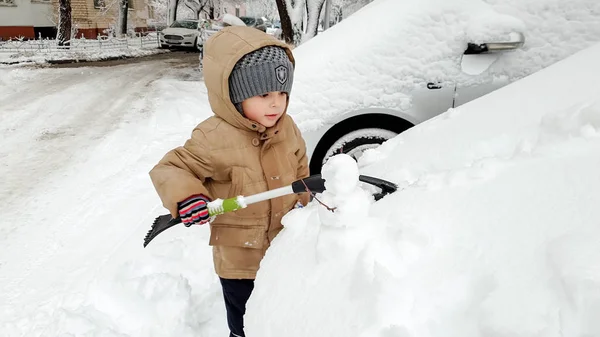 Close up photo of cute smiling boy in warm gloves hat and jacket helping to clean up snow covered parents red car after blizzard using big brush. В холодное снежное зимнее утро — стоковое фото