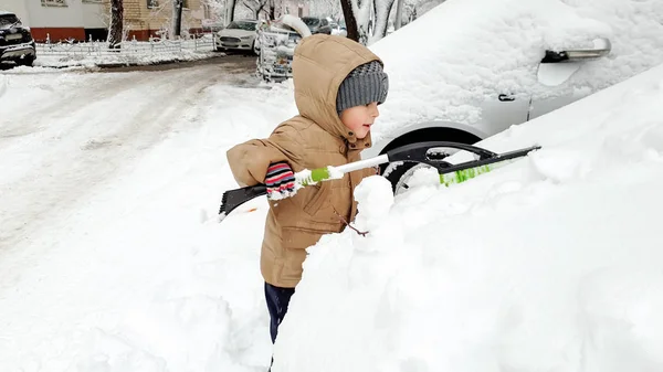 Zbliżenie obraz cute uśmiechnięty chłopak w rękawice kapelusz i kurtka pomaga oczyścić śnieg pokryte rodzice czerwony samochód po Blizzard za pomocą Big Brush. W zimny śnieżny zimowy poranek — Zdjęcie stockowe