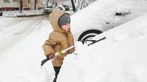 Feche o retrato do rapaz sorridente encantador em chapéu de luvas e jaqueta que ajuda a limpar o carro de pais coberto de neve depois da nevasca usando a grande escova. Na manhã fria de inverno nevado — Fotografia de Stock