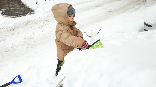 Крупный план улыбающегося мальчика в перчатках и куртке, помогающего убирать снежную машину родителей после метели большой кистью. В холодное снежное зимнее утро — стоковое фото