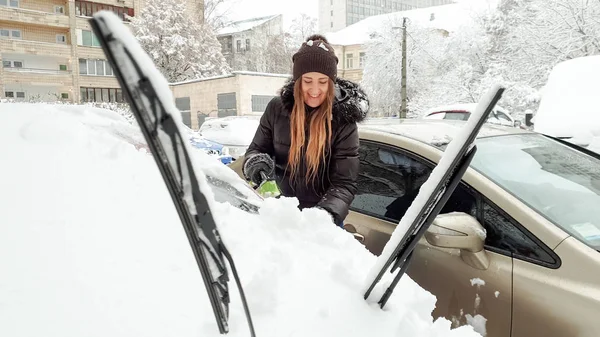 Closeup image of happy lady in jacket and jeans trying to clean up snow covered red auto by brush. Scraping the windshield and wipers of snow at the early snowy winter morning — Stock Photo, Image