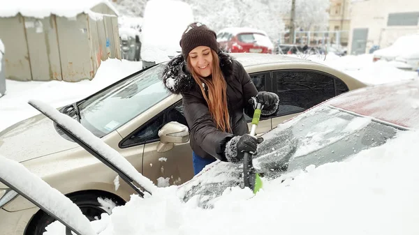 Zavřete fotografii usměvavé ženy ve svěrací kazajce a džínách, které se snaží vyčistit sníh zakrytý červeným automatem. Seškrabání čelního skla a stěračů sněhu pomocí škrabky na časné zasněžené zimní ráno — Stock fotografie