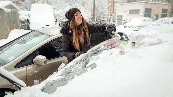 Närbild skott av leende blond dam i jacka hatt och jeans försöker städa upp snötäckta röda Auto med pensel. Skrapa vindrutan och vindrutetorkare med skrapan på den tidiga snöiga vintern — Stockfoto