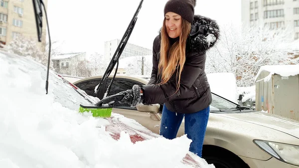 Closeup portrait of young beautiful smiling blond woman in brown jacket hat gloves and blue jeans trying to clean up snow covered red auto by brush after snow storm. Scraping the windshield and wipers — Stock Photo, Image