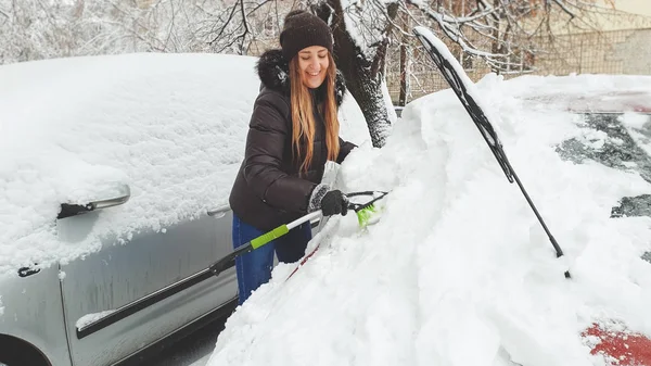 Närbild skott av leende kvinna i jacka handskar och jeans försöker städa upp snötäckta röd Auto med pensel efter Snö Storm. Skrapning av vindrutan och vindrutetorkare med skrapan i början av — Stockfoto