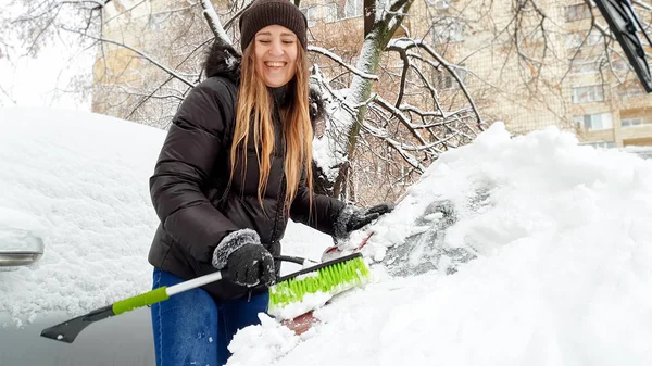 De cerca la imagen de la chica feliz en la chaqueta tratando de limpiar la nieve cubierta de auto rojo por el pincel verde después de la tormenta de nieve. Raspando el parabrisas y limpiaparabrisas de nieve usando el rascador a principios del invierno nevado —  Fotos de Stock