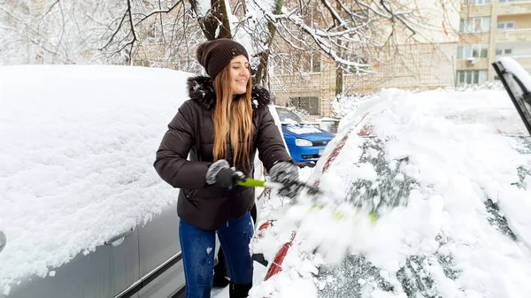 Primo piano colpo di ragazza felice in giacca cercando di ripulire la neve coperto auto rossa da spazzola verde dopo bufera di neve. Raschiare il parabrezza e i tergicristalli di neve usando il raschietto all'inizio dell'inverno nevoso — Foto Stock