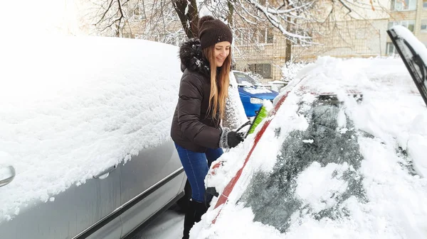 Close up shot of girl trying to clean up snow covered red auto by green brush after blizzard. Scraping the windshield and wipers of snow using the scraper at the early snowy winter morning — Stock Photo, Image