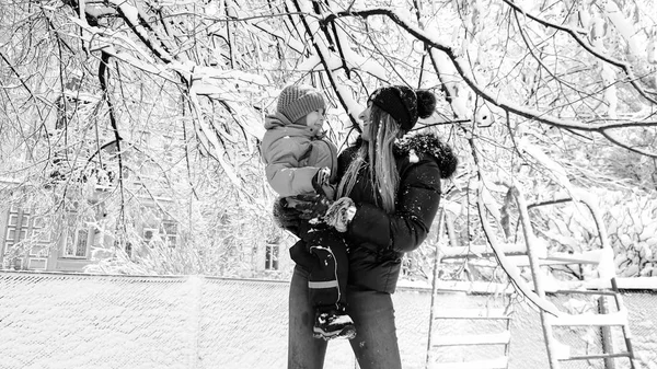 Monohrome retrato da mãe com seu filho bonito em casaco e chapéu brincando com a árvore coberta de neve no playground no parque — Fotografia de Stock