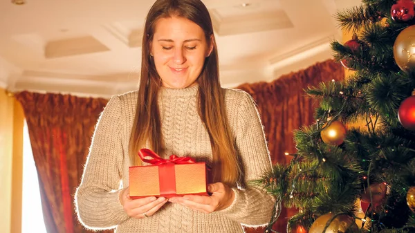 Toned portrait of happy smiling young woman standing with gift box next to Christmas tree in living room — ストック写真