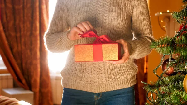 Closeup image of young woman holding box with Christmas present and pulling ribbon bow to open the gift — ストック写真