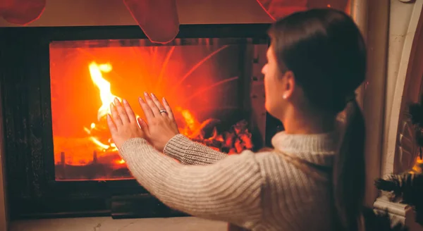 Toned image of beautiful woman feeling cold sitting by the burning fireplace at living room — Stock Photo, Image