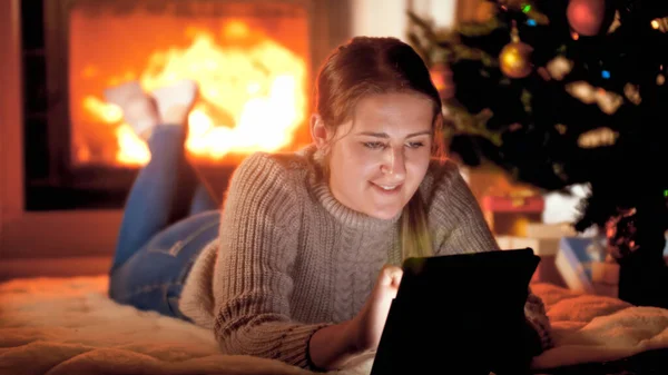 Retrato de una hermosa mujer sonriente en suéter tumbado junto a la chimenea bajo el árbol de Navidad y utilizando una tableta digital — Foto de Stock