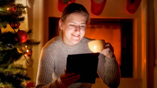 Retrato de uma jovem feliz usando tablet digital e bebendo chá enquanto se senta na lareira e na árvore de Natal — Fotografia de Stock