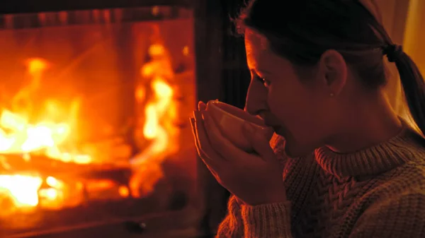 Closeup toned image of young woman drinking tea at night while sitting by the burning fireplace at house — Stock Photo, Image