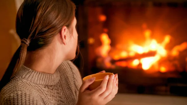 Portrait of woman in woolen sweater holding cup of hot tea and looking at burning firepalce — Stock Photo, Image