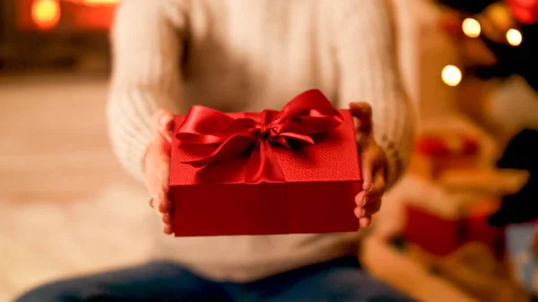 Foto de primer plano del joven owman sentado en el suelo junto al árbol de Navidad y mostrando la caja de regalo roja en la cámara —  Fotos de Stock